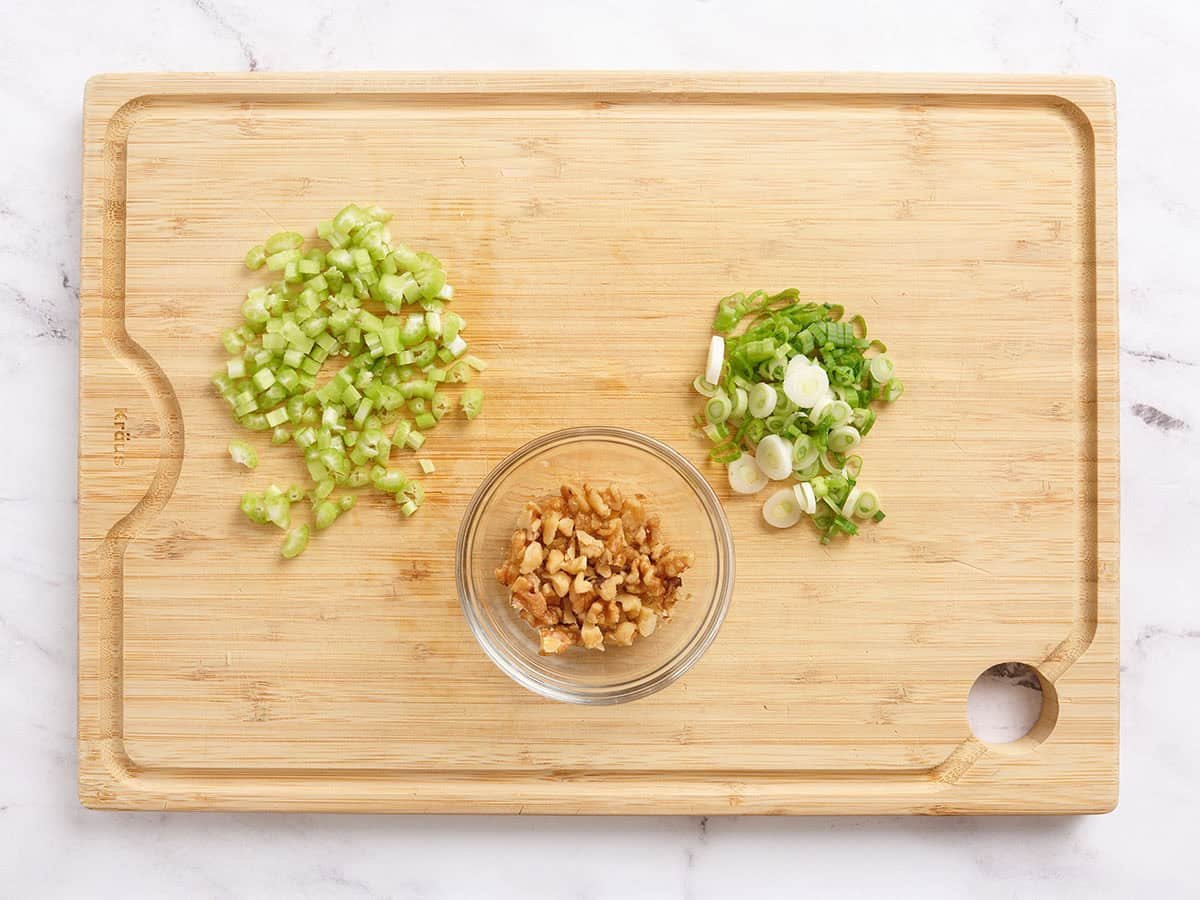 Diced green onions, chopped walnuts, and diced celery on a wooden cutting board.