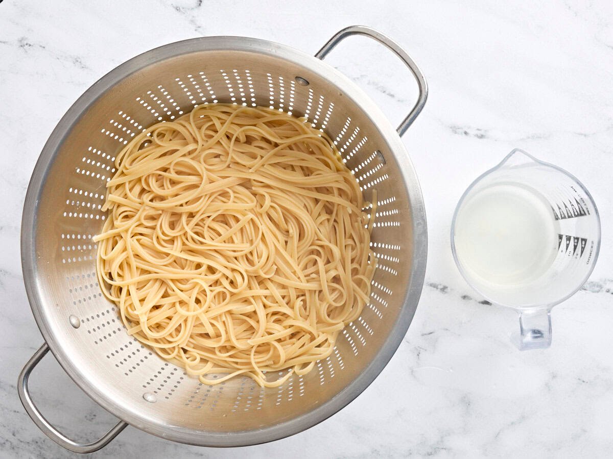 Boiled Fettuccine in a colander, next to a glass jug of reserved pasta water.