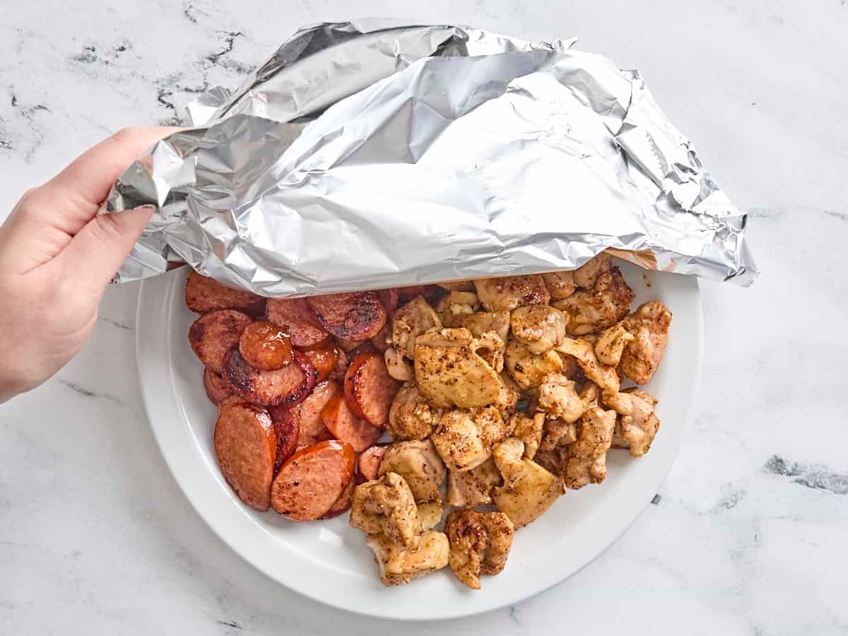 Browned chicken and sausage on a plate being covered with foil.