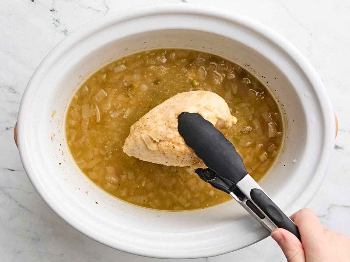 A chicken breast being removed with tongs from a crockpot of slow cooker white chicken chili.