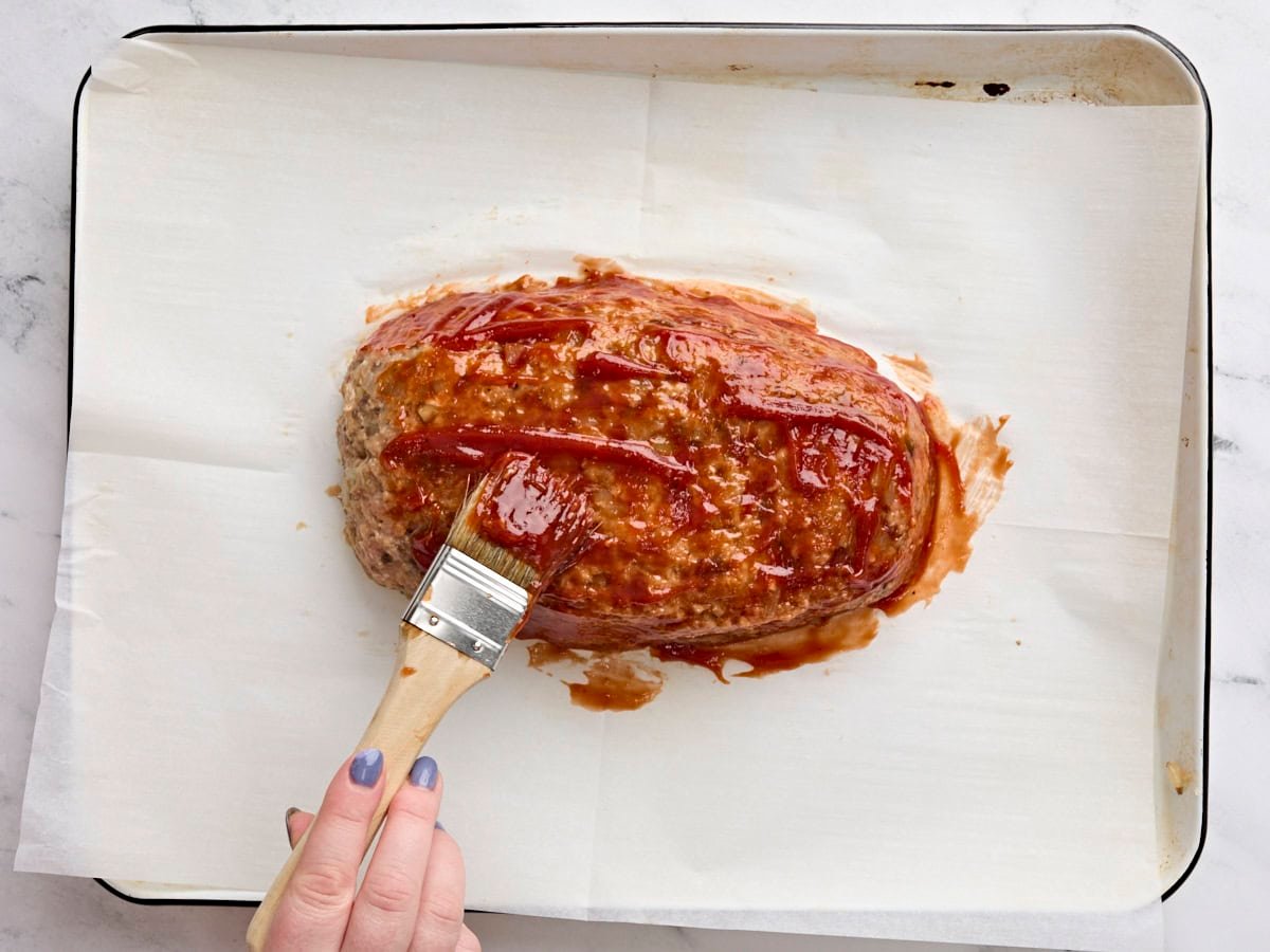 A turkey mushroom meatloaf being brushed with BBQ sauce before being baked on a parchment lined baking sheet.