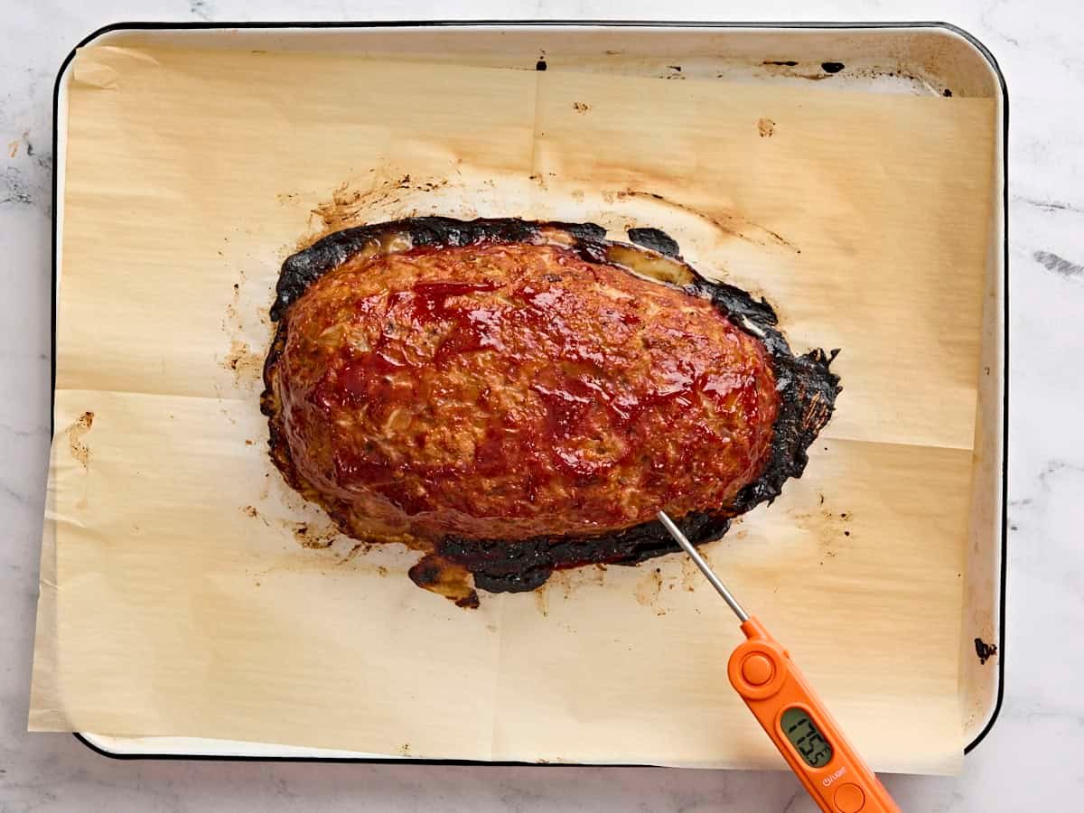 A meat thermometer checking the internal temperature of a freshly baked turkey meatloaf on a parchment lined baking sheet.