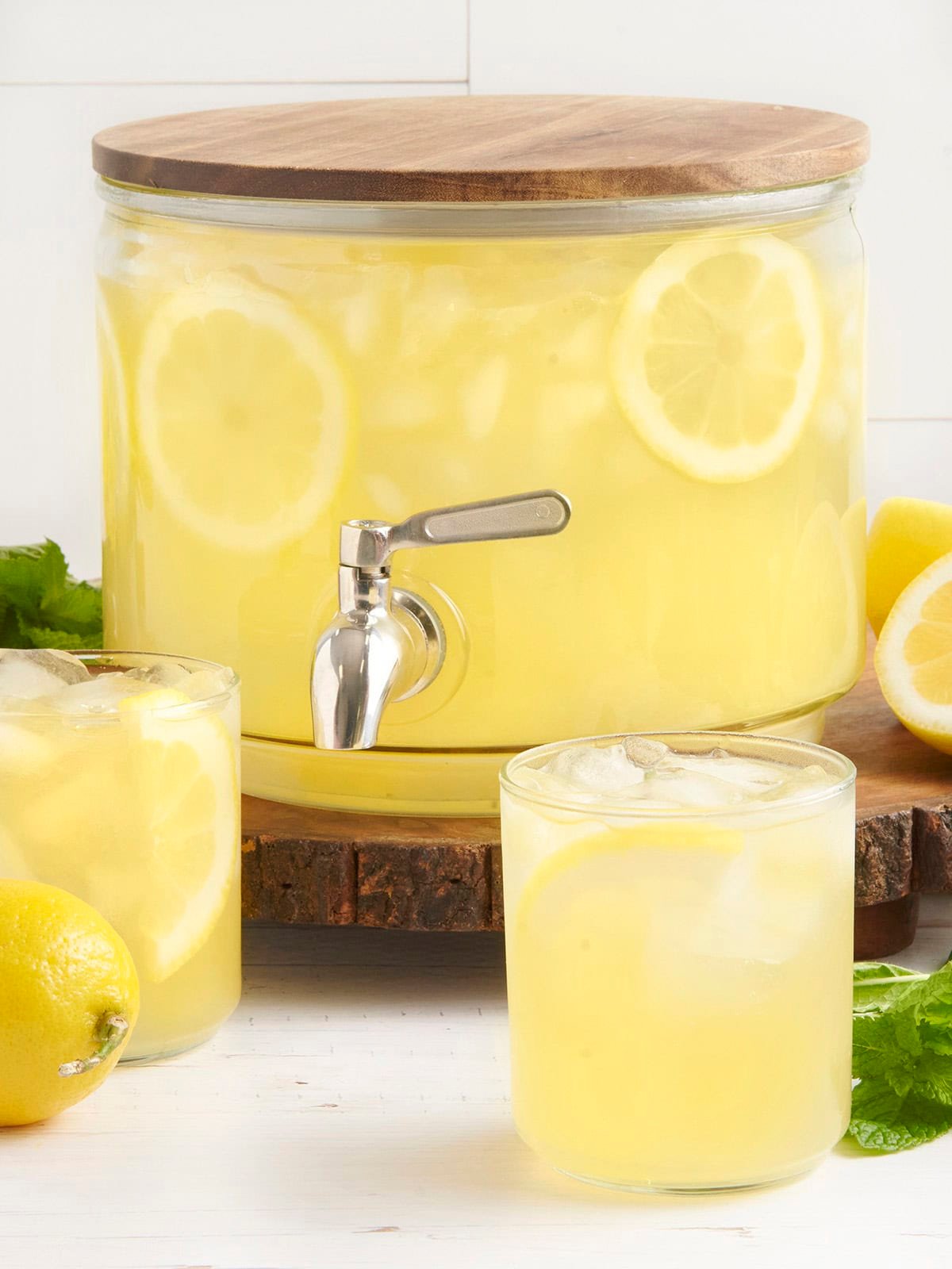 A drink dispenser with homemade lemonade and two glasses of lemonade in front.